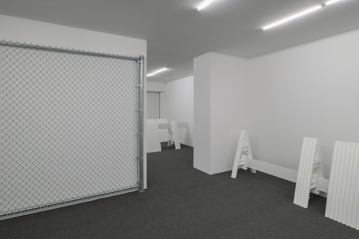 In a room with white walls and gray carpet, a portion of a chain link fence stands against a wall and plastic barricades are placed nearby.
