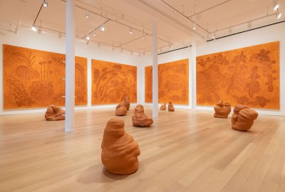Installation view of a gallery showing four large-scale paintings on the wall and several clay-mound-like sculptures on the floor. They all share the same burnt-orange, earthy color palette.