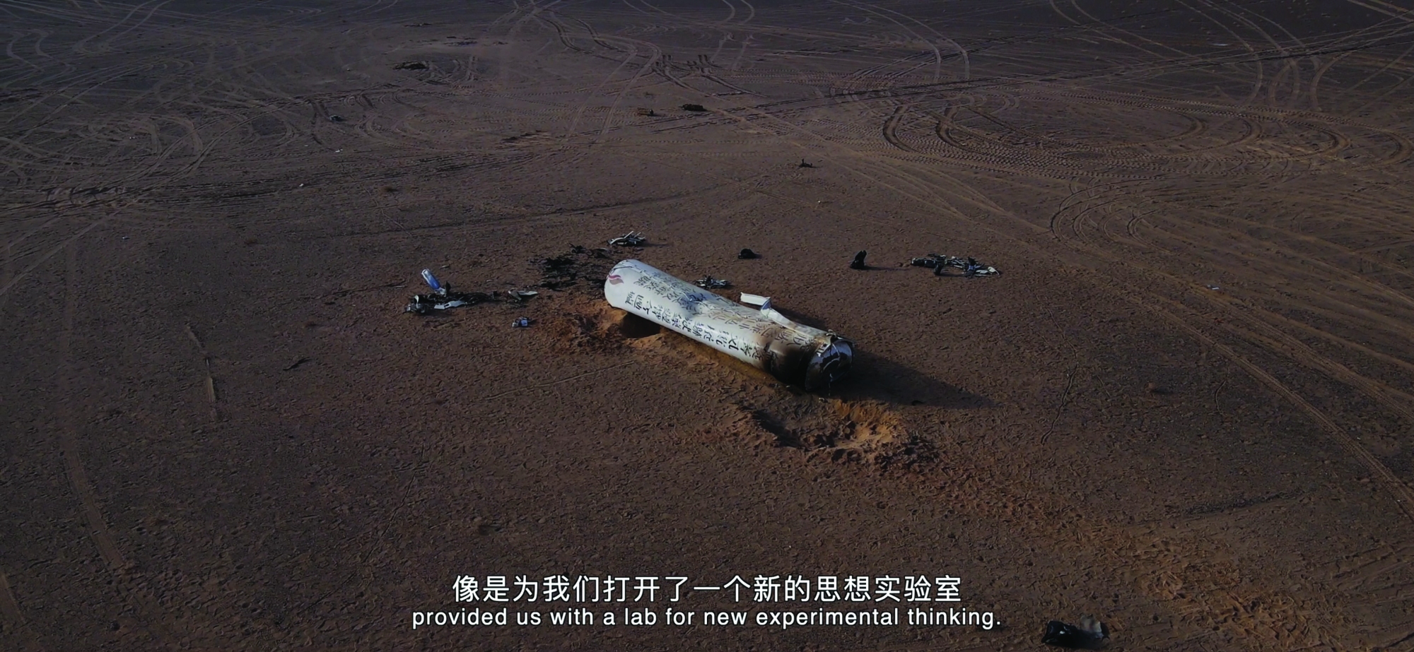 A large tubular white rocket with fake Chinese characteres printed on it has crashed into a vast desert and left an indent.
