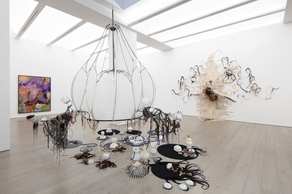 A brightly lit gallery filled with a large installation resembling a fallen chandelier that has hair and other elements affixed to it.
