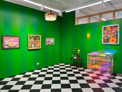 A gallery room with green-striped walls and a black-and-white diamond tile floor. There are paintings on the wall, a chandelier, a table with flowers, and a vitrine with neon squiggles.