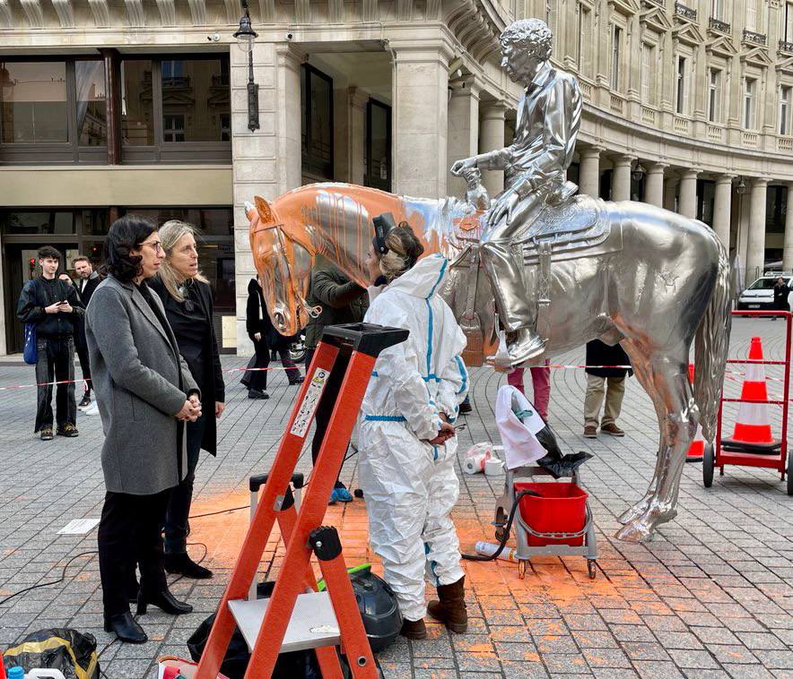 Two white women look at a stainless steel sculpture of a man riding a horse that has been doused in orange paint. A worker in a haz-mat suit stands to the right as cleaning of the sculpture is in progress.
