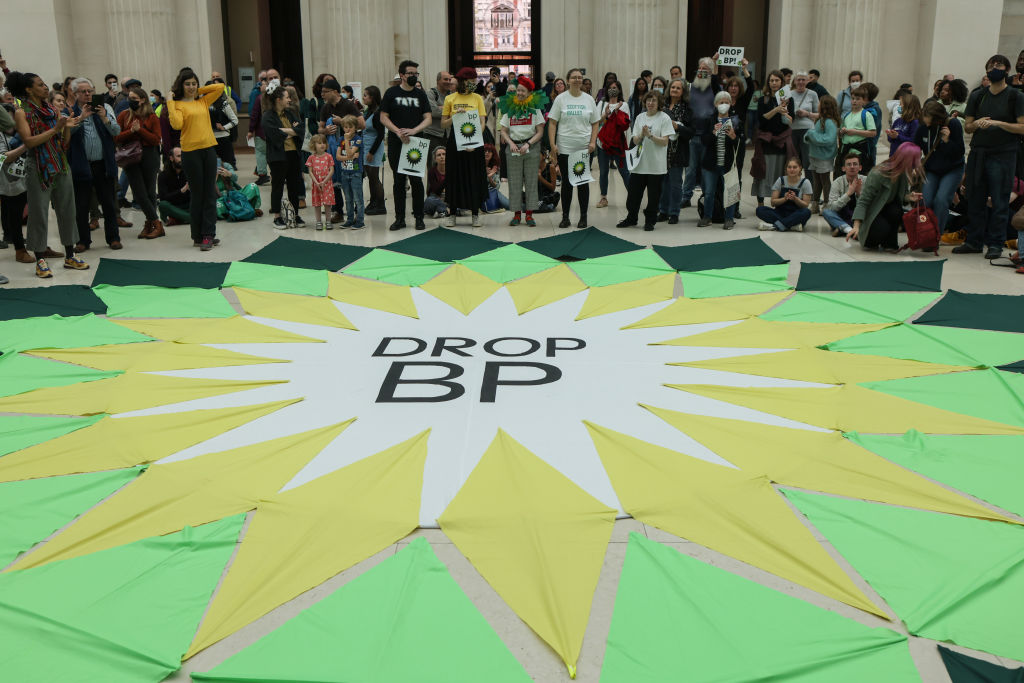 A group of people stand around a bright green and yellow banner that says "Drop BP" in the style of the BP logo, which resembled an angular sun or flower. They are in a large hall.