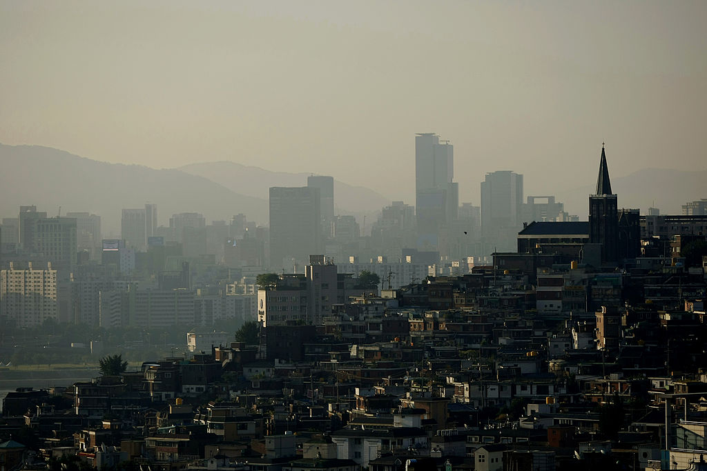 A vast cityscape is seen from the air, with many buildings climbing a steep slope. The air is hazy.