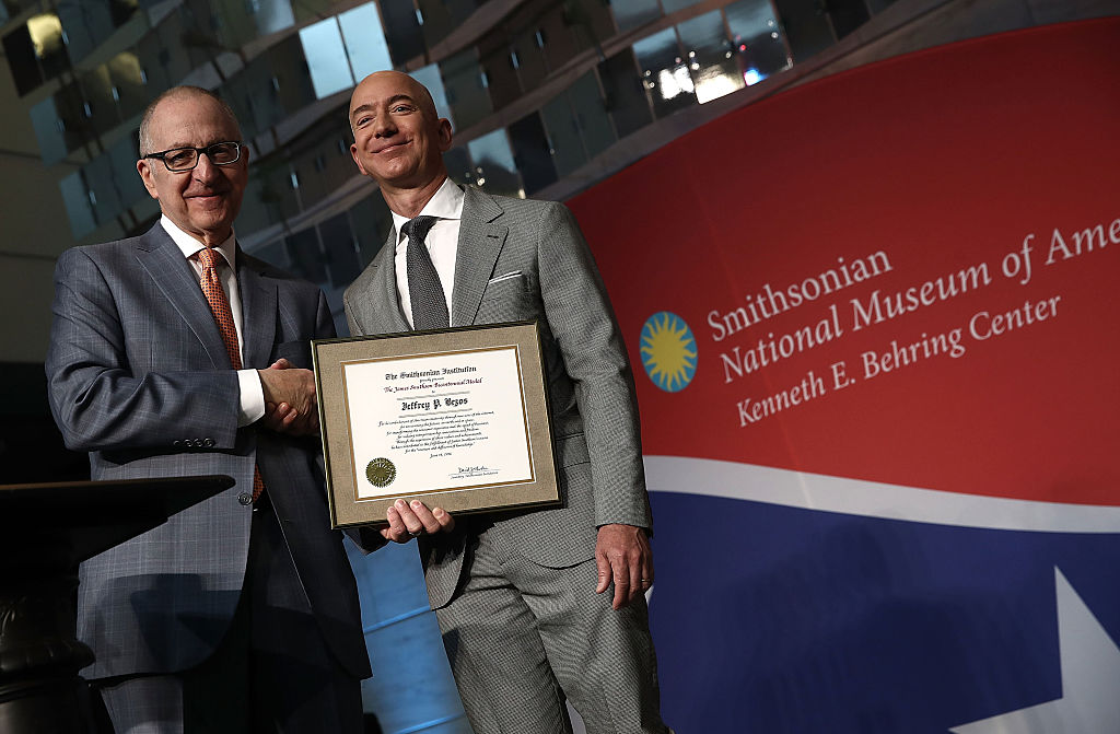 Two men in suits stand on a stage and shake hands. One holds a framed certificate; the other smiles broadly.