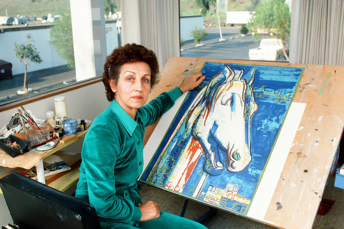 A woman seated a painter's table on which lies a painting of a horse that is half-finished.