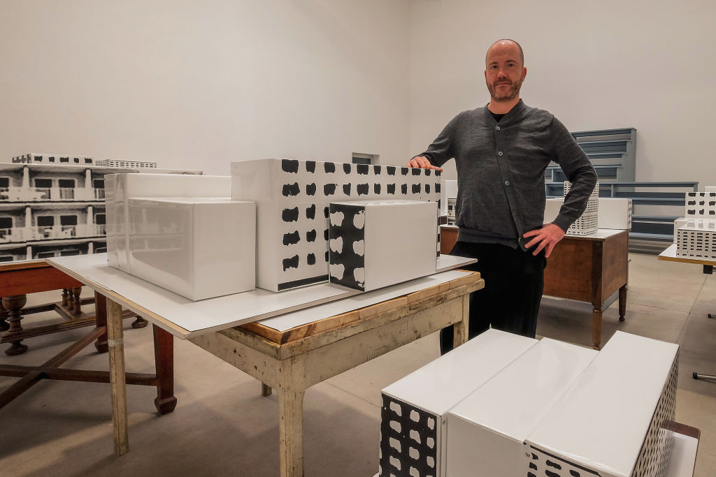 A man stands amid an art show in a white-walled gallery with high ceilings. Tables of various kinds hold what appear to be rudimentary models for buildings.