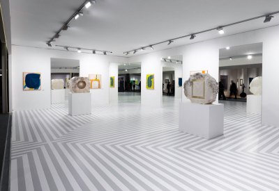 View of an art fair booth with a gray-white chevron pattern on the floor, paintings and mirrors alternating on the walls, and two sculpture on plinths at center.