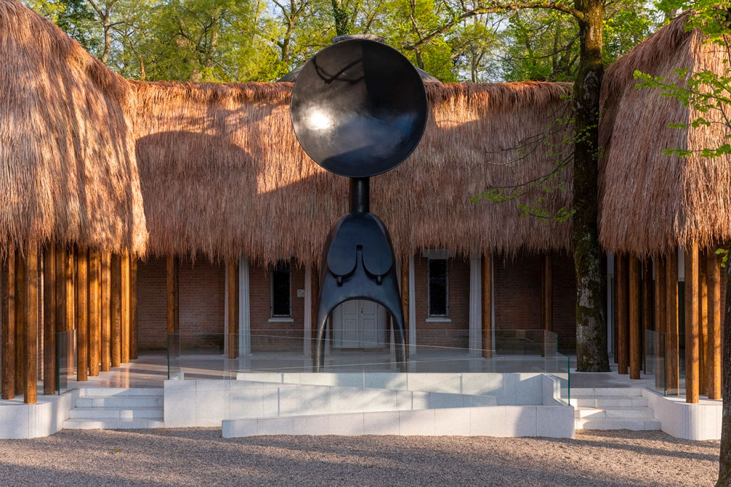 A pavilion with a thatched roof, with a sculpture of a nude femme in its center. The figure's head is replaced with a giant bowl-like form, and its legs appear more like prongs. A ramp leads up to the sculpture.