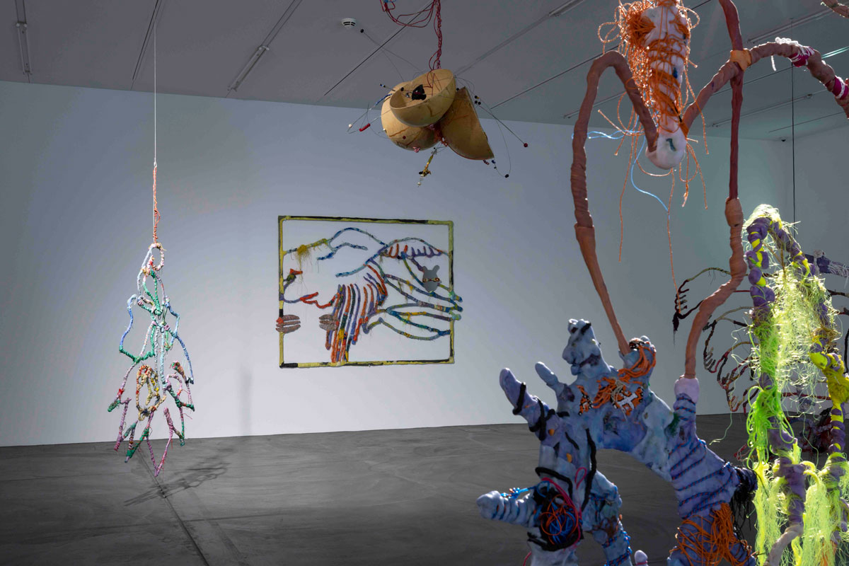 Installation view of a museum exhibition showing various sculptural works suspended from the ceiling. 
