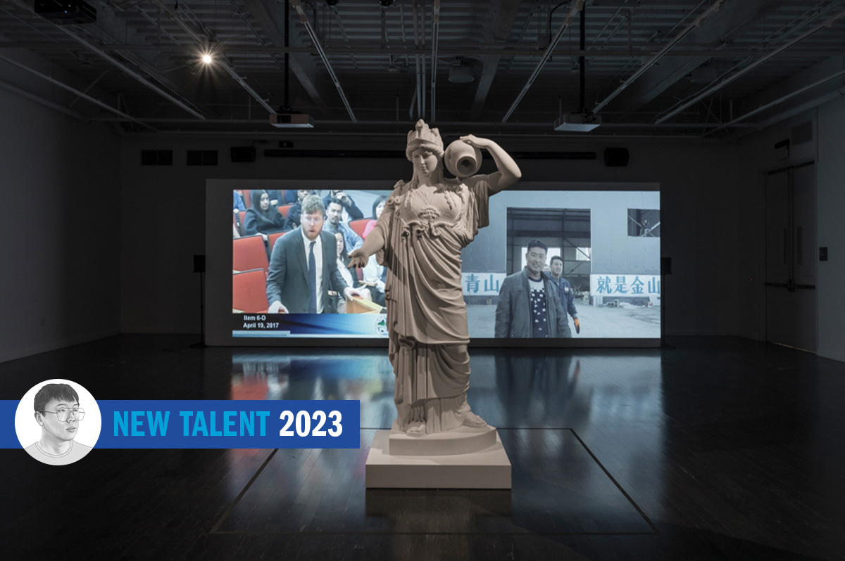 installation shot showing a carved sculpture of a figure in robes in the foreground with a video projection in the background