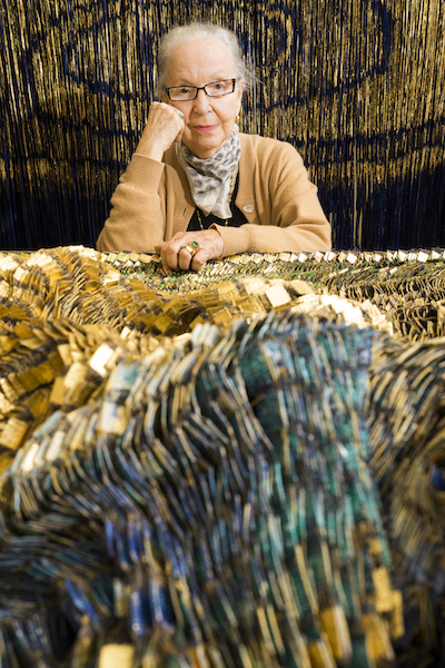 Surrounded by beaded yarn, an elderly lady with glasses sits, with her head propped on one fist, looking straight out at the viewer.