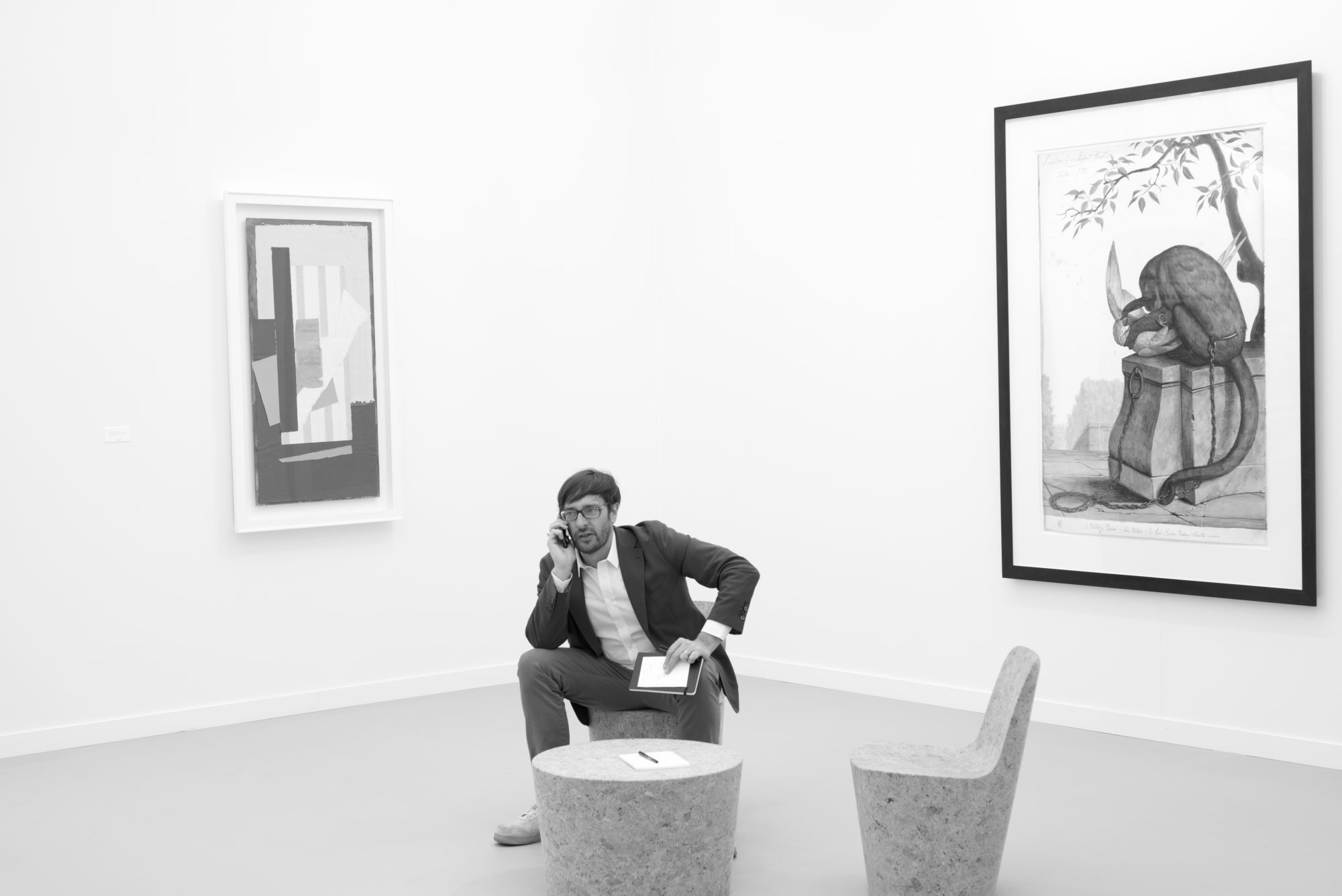 A man sits on a chair between two pieces of art and speaks on the phone.