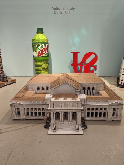 An intricate cardboard sculpture of a building, with a sculpture of a Mountain Dew bottle and "LOVE" sculpture behind it.