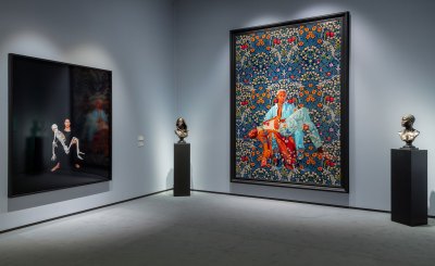 View of an art fair booth with a photograph of a woman holding a skeleton at left. At right a painting of a Black man holding another Black man on a floral background. The painting is flanked by two bronze sculptures.