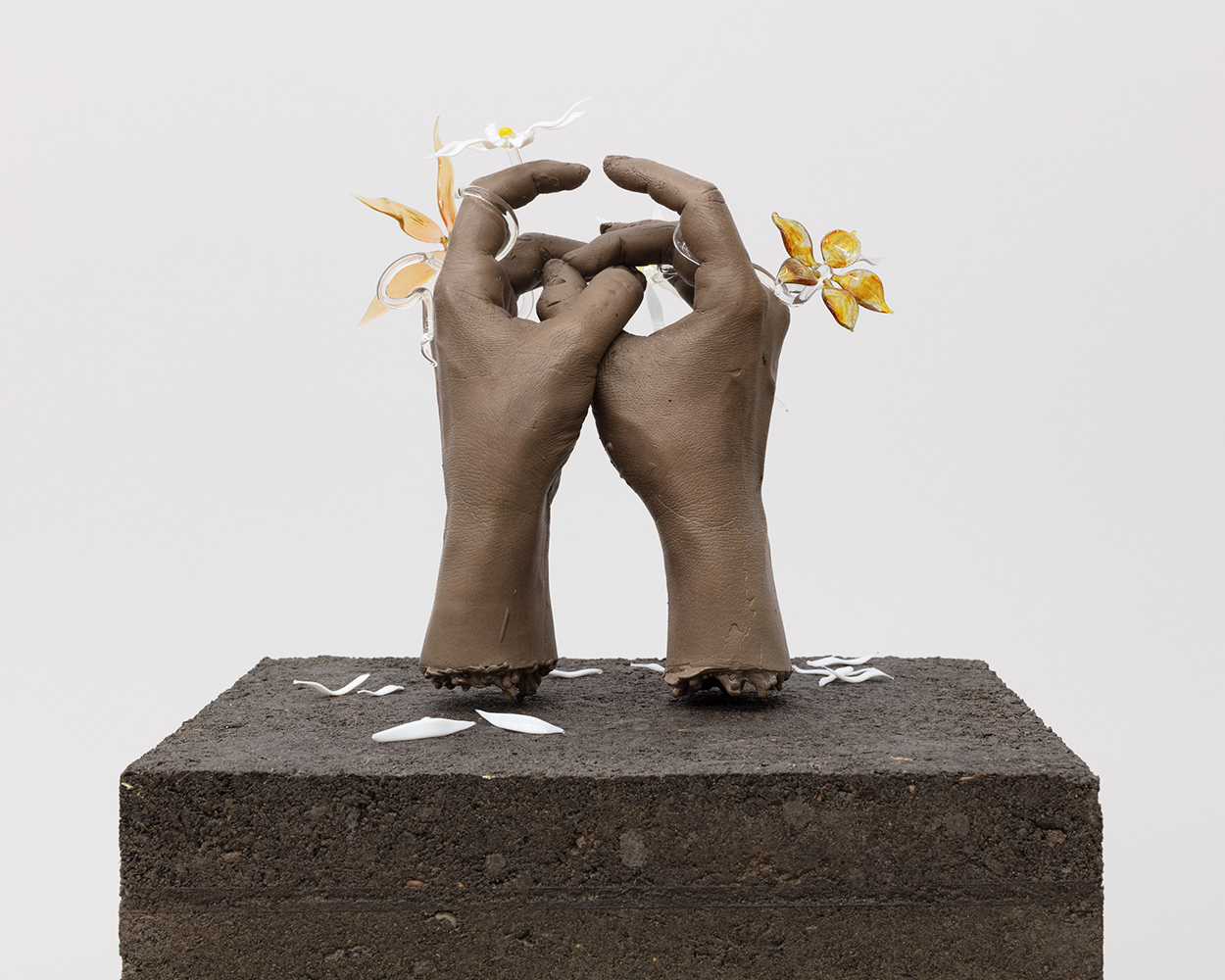 On a plinth amde of dirt, two bronze hands create interlinking circles with their middle fingers and thumbs. Delicate glass flowers encircle both, and leave petals in the dirt.