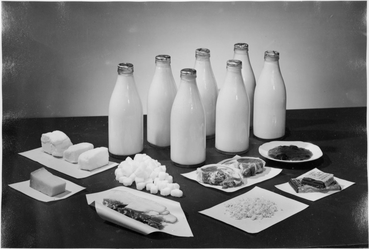 A black-and-white photo showing 7 bottles of milk and other foods.